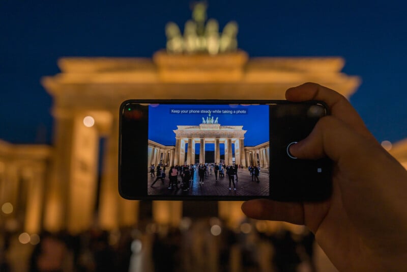 A person holds a smartphone to take a picture of an illuminated historical building at night. The building is glowing with warm lights, and people are gathered in front of it.