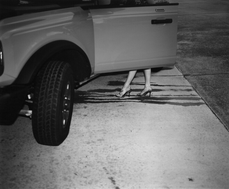 Black and white image of a person stepping out of a car. Only the lower half is visible, wearing high heels. The car door is open against a concrete surface with a liquid spill.