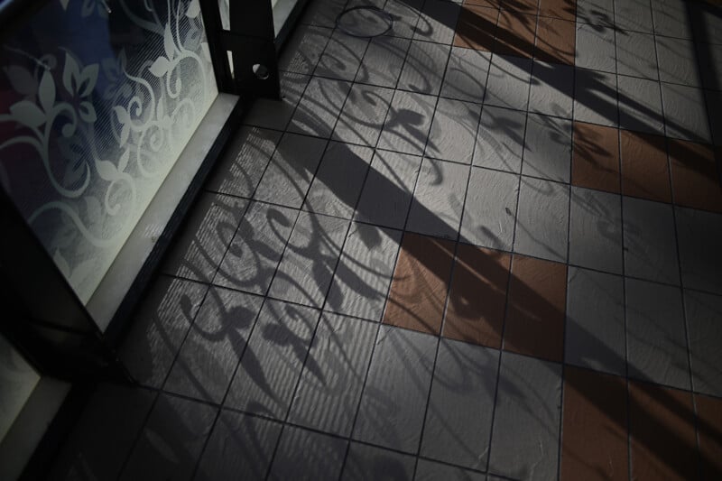 Sunlight casts intricate shadow patterns from a decorative glass window onto a tiled floor. The tiles are alternating shades of gray and brown, creating a contrast with the flowing, abstract shapes of the shadows.