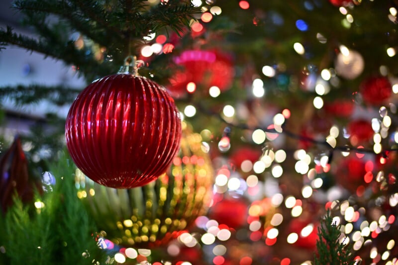 A close-up of a Christmas tree decorated with a shiny red bauble and blurred multicolored lights in the background, creating a festive, sparkling atmosphere.