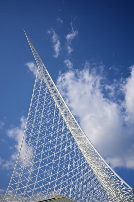 A modern, white steel architectural structure resembling a sail against a bright blue sky with scattered clouds. The intricate lattice design creates an airy and dynamic visual.