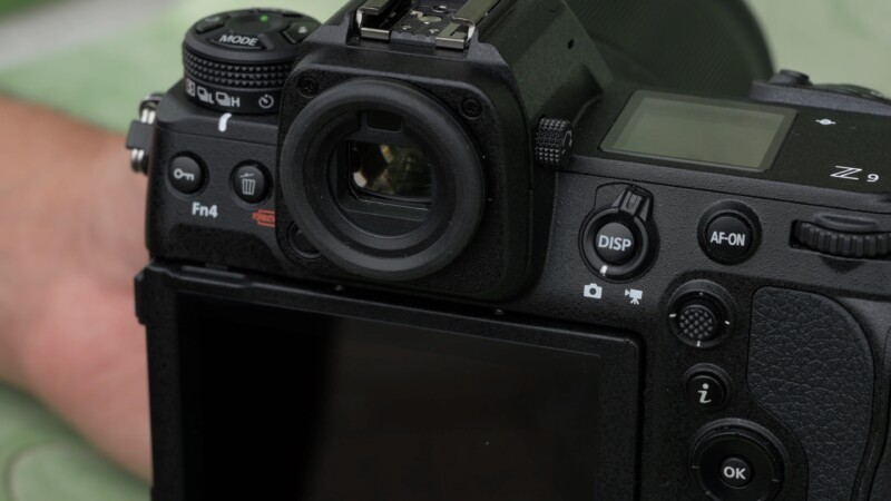 Close-up of a black digital camera showing buttons and dials on the back, including the viewfinder, DISP, and AF-ON buttons. The camera is positioned on a green surface.