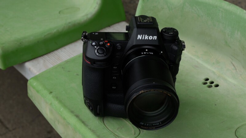 A Nikon camera with a lens is placed on a green plastic chair. The camera has a black body, and various buttons and a display screen are visible. The chair has a textured surface with circular aperture-like patterns.