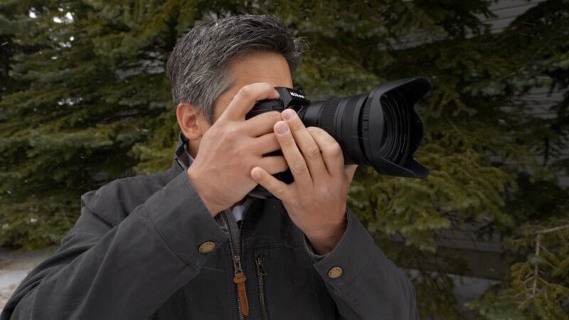 A person with gray hair is holding and looking through a large camera with both hands. They are wearing a dark jacket and standing outdoors in front of evergreen trees.