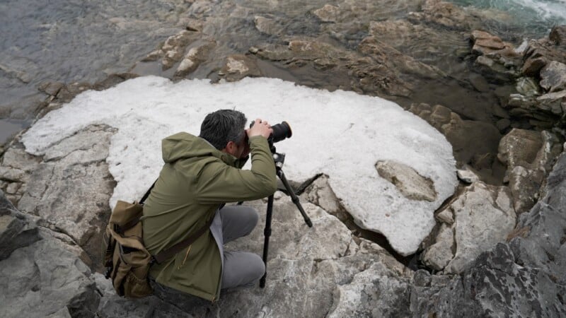 A person in a green jacket kneels on rocky terrain, using a camera on a tripod to photograph a patch of melting ice near a body of water. They carry a brown bag, and the scene suggests a rugged, natural environment.