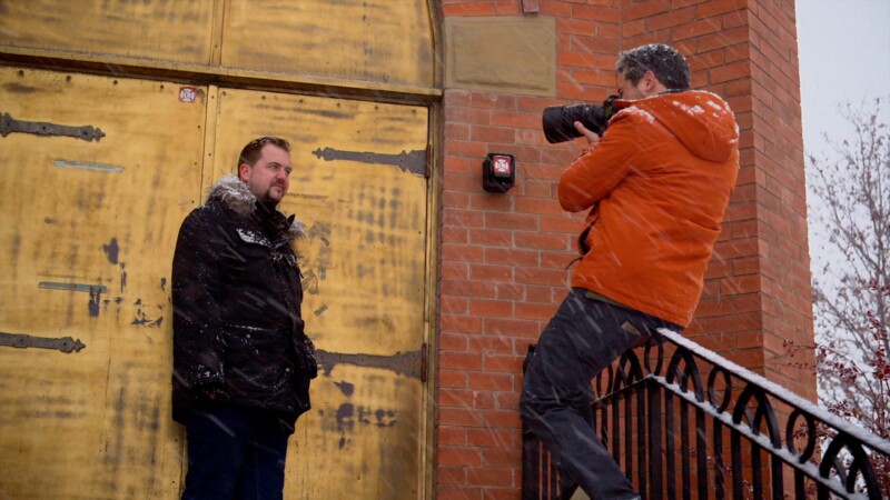 A photographer in an orange jacket and snow-covered hair crouches on stairs, taking a picture of a man in a black coat standing against a large, aged gold door with decorative elements. Snowflakes fall around them.