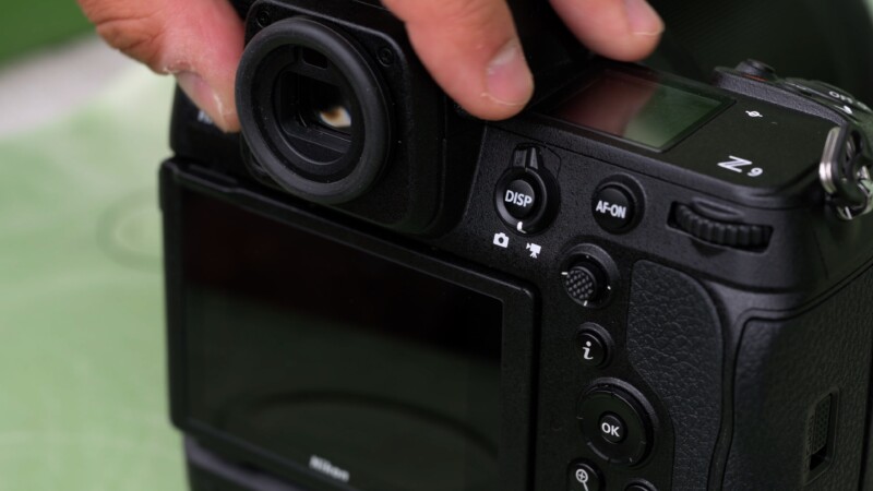 Close-up of a person holding a black camera, adjusting buttons near the viewfinder. The camera's display screen and various control buttons are visible.